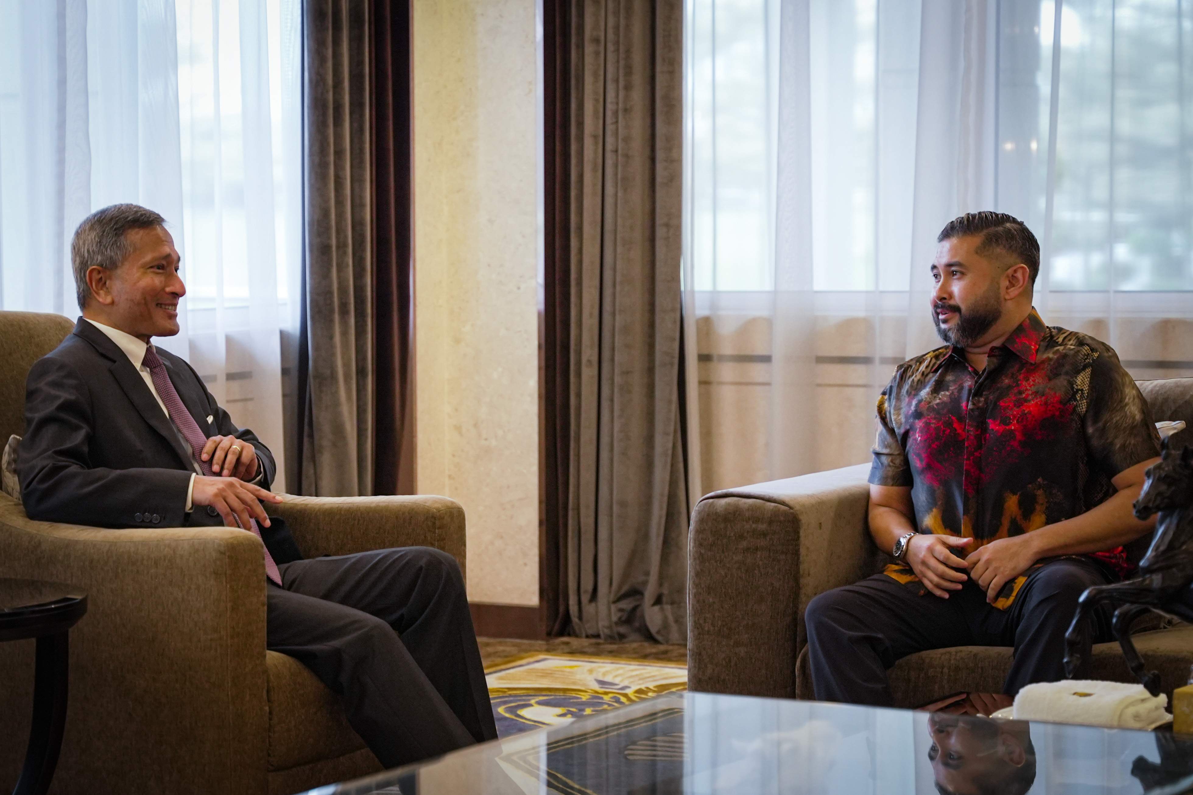 Two men in armchairs facing each other in a room with curtains and a glass table.